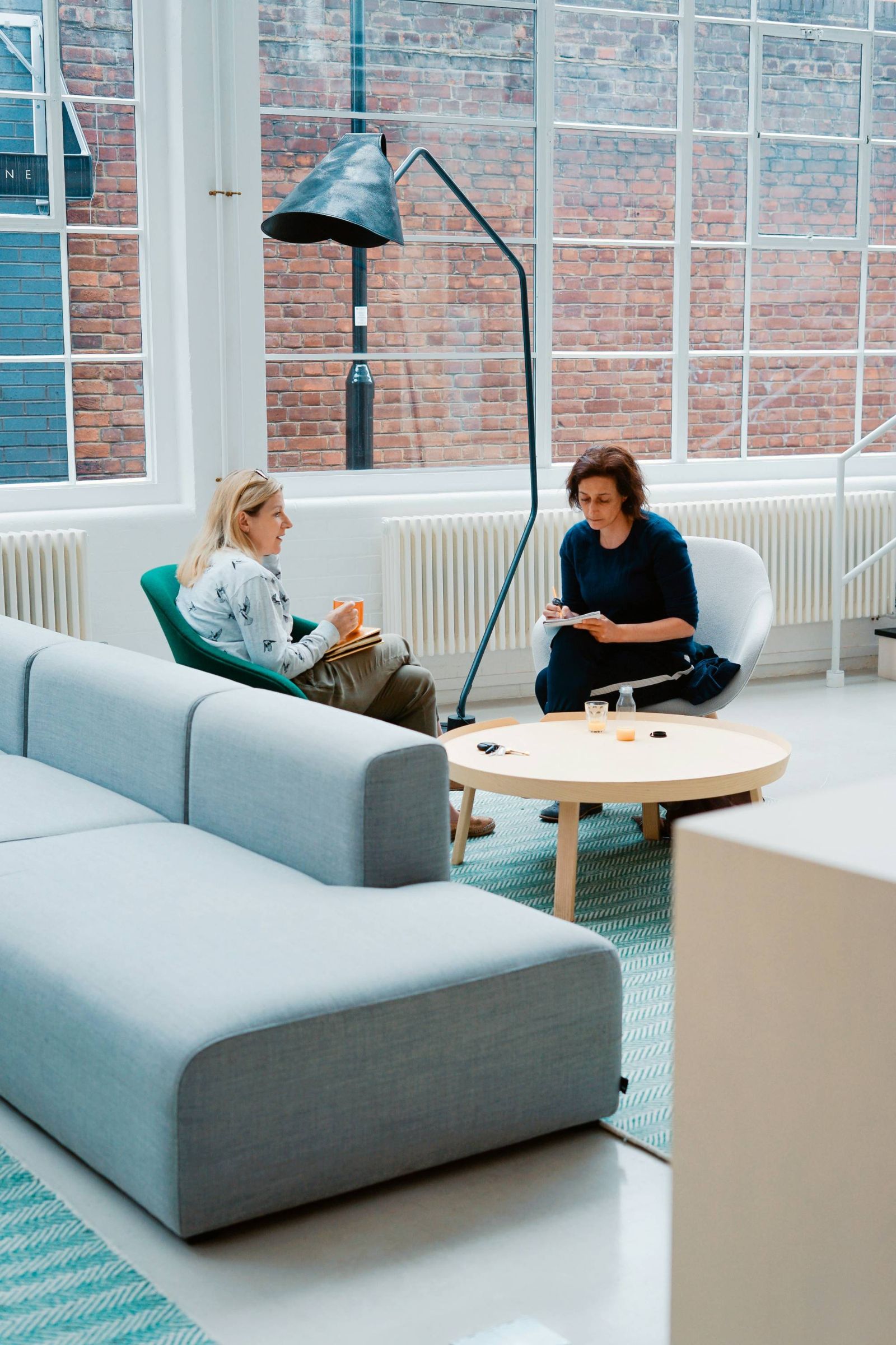 Two people talking on a sofa beside tall windows in a modern office