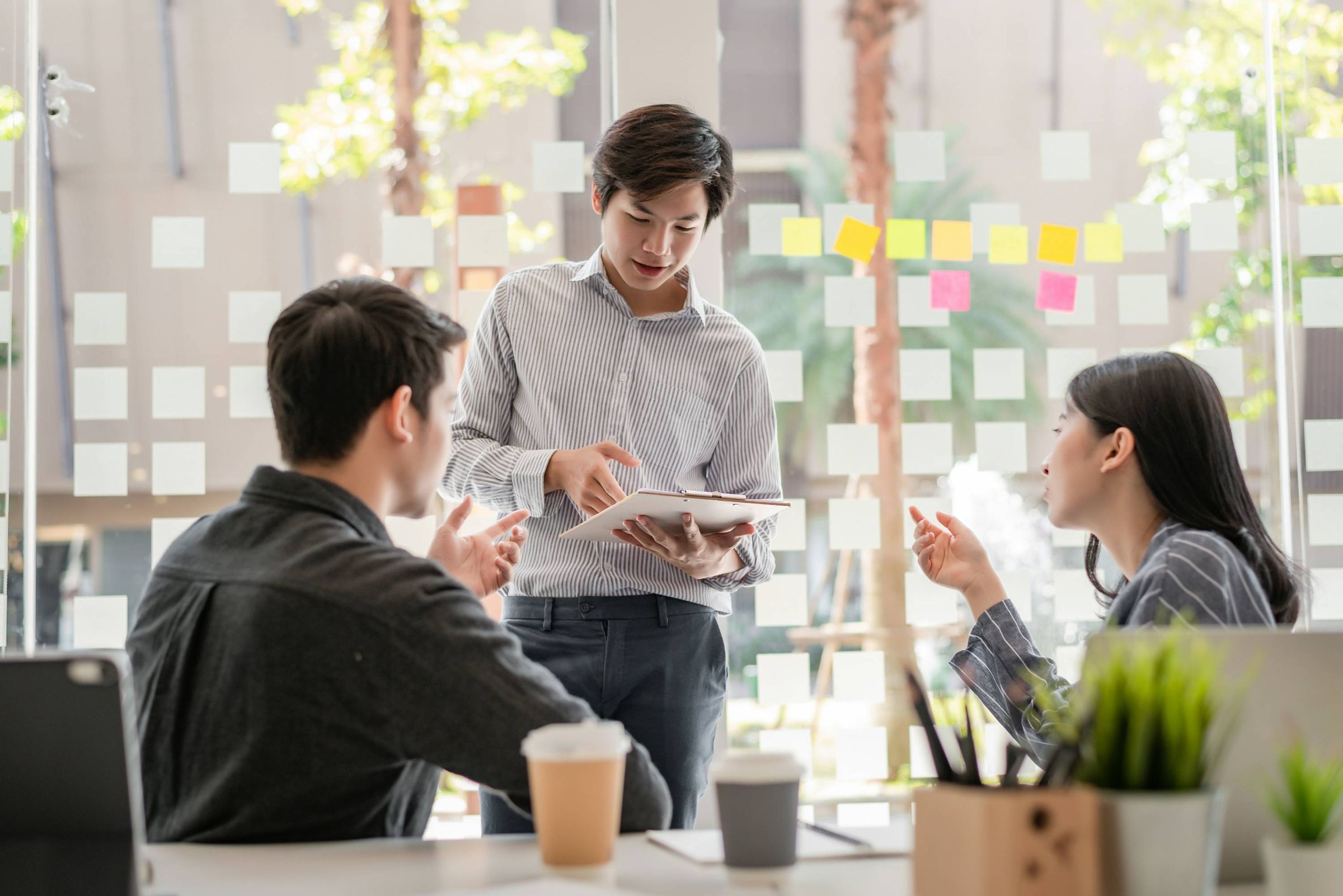 Colleagues discussing a document at a desk in front of a glass wall of sticky notes